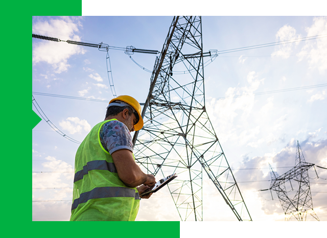 A man with a hard hat and safety vest stands near an electricity tower.